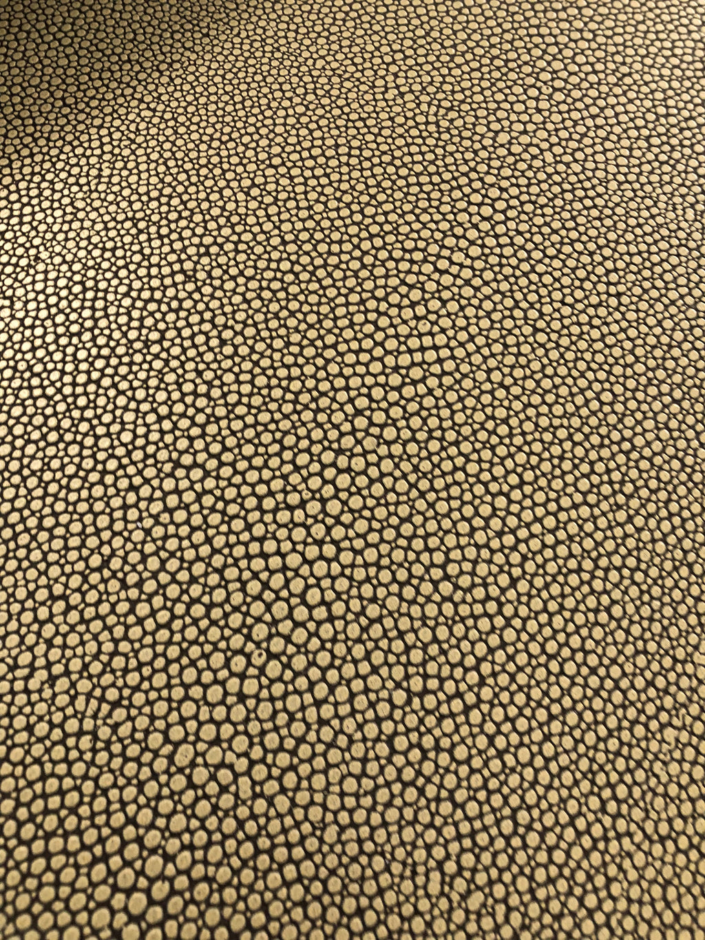 A close-up of a textured material featuring a repeating pebble or reptile-scale pattern in black on a warm tan or beige background.