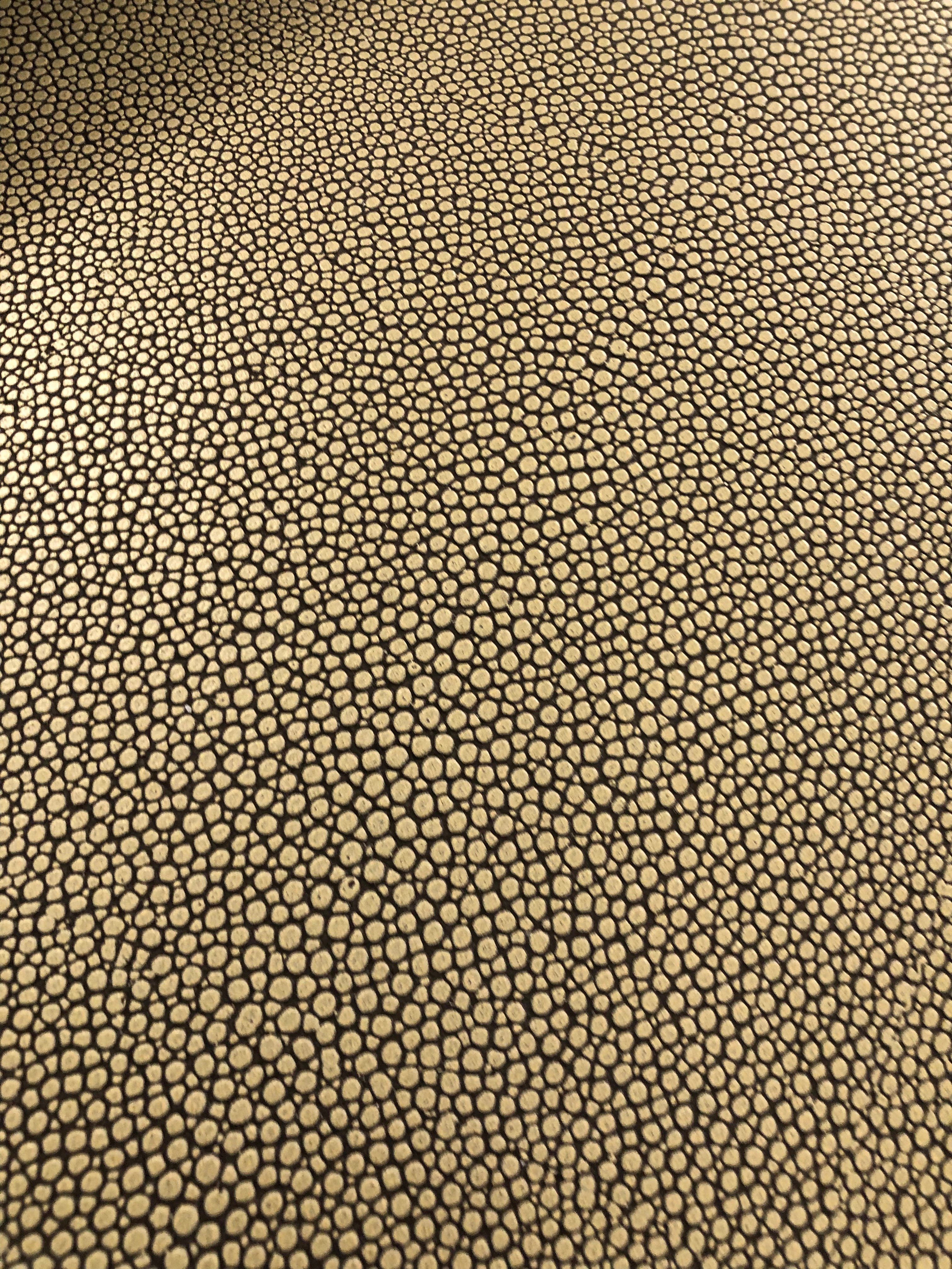A close-up of a textured material featuring a repeating pebble or reptile-scale pattern in black on a warm tan or beige background.