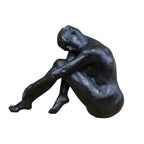 A dark bronze-finished sculpture of a seated nude human figure with knees drawn up and head resting on arms, displayed on a light wood surface against a blurred light brick wall background.