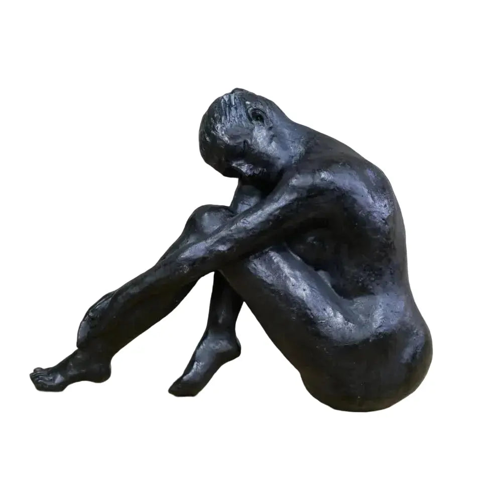 A dark bronze-finished sculpture of a seated nude human figure with knees drawn up and head resting on arms, displayed on a light wood surface against a blurred light brick wall background.