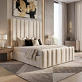 A modern bedroom featuring a cream upholstered bed with vertical channel tufting, set against a textured gray wall with abstract black and gold wall art. The room includes light wood flooring, matching geometric area rugs, bedside tables with lit lamps, and a large window with sheer curtains.