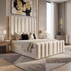 A modern bedroom featuring a cream upholstered bed with vertical channel tufting, set against a textured gray wall with abstract black and gold wall art. The room includes light wood flooring, matching geometric area rugs, bedside tables with lit lamps, and a large window with sheer curtains.