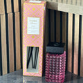 Pink textured diffuser bottle with black cap next to its packaging on a wooden surface.