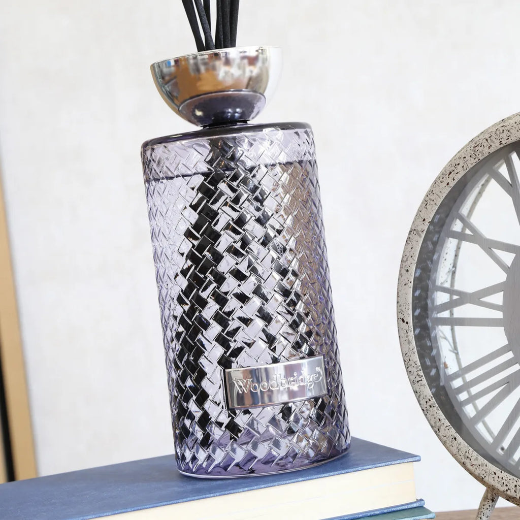 A Woodage reed diffuser with a textured, woven-look metallic purple glass body, resting on a blue book next to a clock.